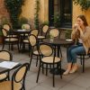 Super-Seat | A woman sits alone at a café table on a Chopin Noir Thonet (terrace) chair with wooden frame, metal terrace tables, glass of water; yellow walls, green plants. | Hospitality Furniture