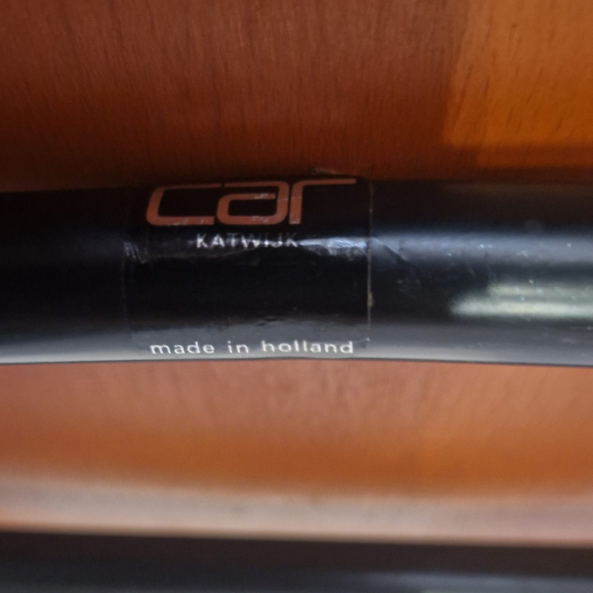 Super-Seat | A close-up of a black tube with two labels—one reading "Car Katwijk," the other "made in holland"—set against a wooden background, echoing the Dutch craftsmanship of Car Katwijk Dutch stackable design chairs. | Zwarte metalen buis, gelabeld, tegen houten achtergrond; typisch voor Nederlands design van stapelbare stoelen. | Horecameubilair