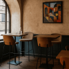 Super-Seat | A small café corner with two wooden tables, several New York design bar stools wood velour Mocha and wooden chairs, a glass bottle and glass on one table, a large arched window on the left and a colorful abstract painting on the wall. | Wooden tables, mocha velour bar stools with wooden legs, wooden chairs; warm tones and glass decor. | Hospitality Furniture
